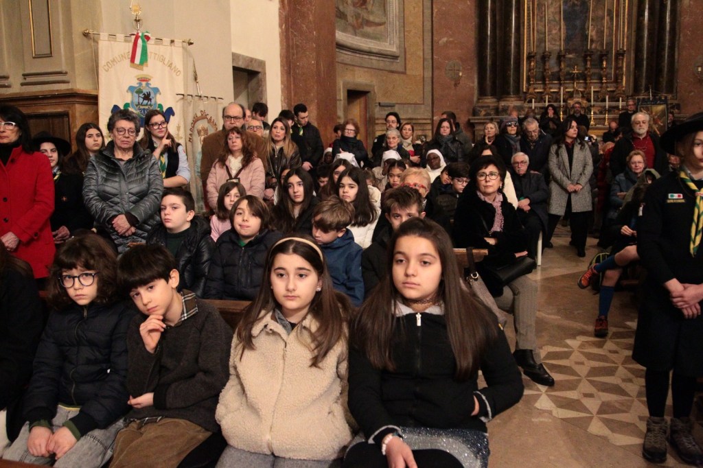 A crowd of children and adults seated in a church, some facing forward and others looking around. The atmosphere appears solemn, and there are various clothing styles represented. A flag is visible in the background.