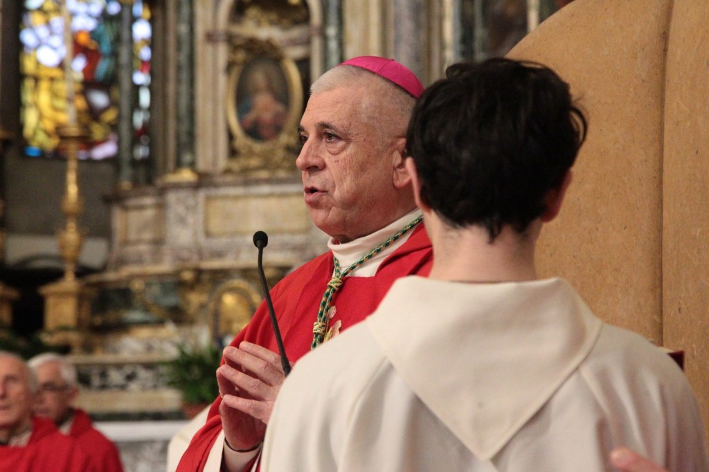 A bishop in red vestments speaks at a podium, addressing a person in white robes in a cathedral setting.