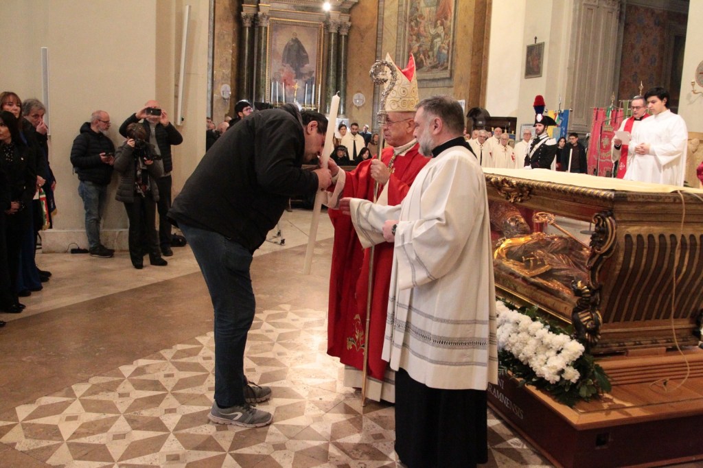 A man leaning forward to kiss the hand of a dignitary during a religious ceremony in a church, with attendees in the background and a decorated altar in the foreground.