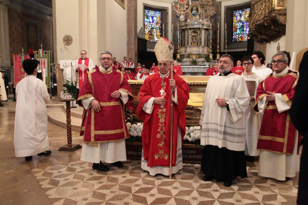 A group of clergy members in red and white vestments stands in front of an altar in a church. The central figure wears a bishop's mitre and holds a staff. The background features decorative stained glass windows and additional clergy in red robes.