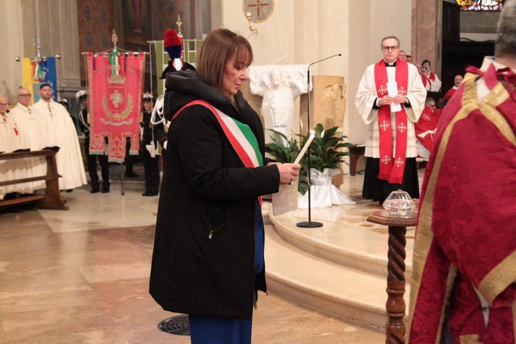 A woman wearing a black coat and a ceremonial sash holds a candle during a religious ceremony, surrounded by people in formal attire and flags in a church setting.