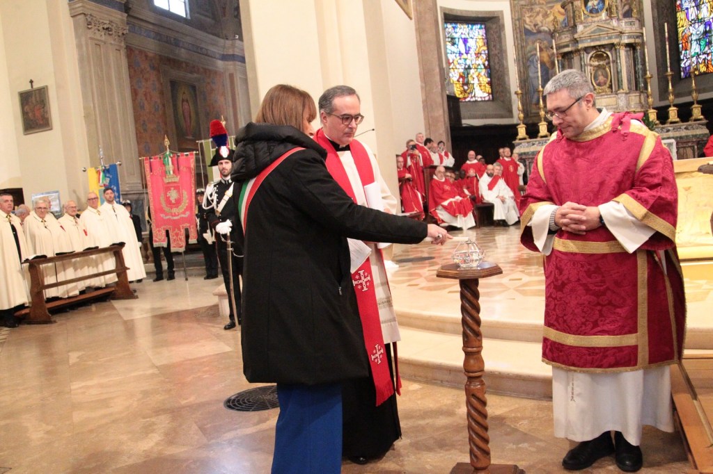 A ceremonial event in a church setting, featuring a woman in formal attire placing a glass container on a pedestal while two men in religious vestments observe. The background includes attendees in ceremonial garments and colorful banners.