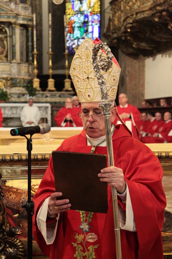 A clergyman in red vestments reading from a book while holding a bishop's crosier, with a microphone in front of him and a congregation in red robes in the background.