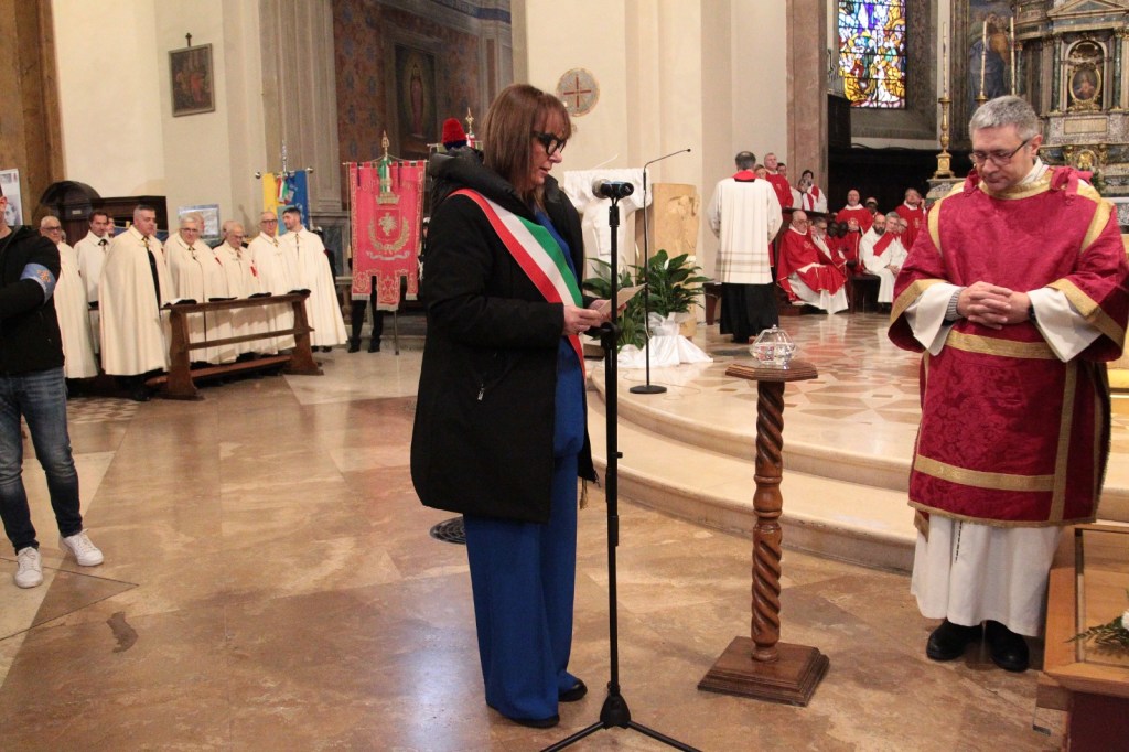 A woman in formal attire stands at a microphone, reading from a paper, while a priest in red robes bows his head beside her. In the background, a group of men in white cloaks stands, and additional clergy in red can be seen in the far background of a church setting.