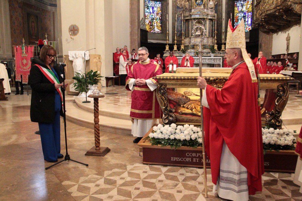 A woman with a microphone reading from a script stands next to a man in a red robe, while another man in a bishop's attire holding a staff looks on. The scene is set in a church with religious figures and decor in the background.