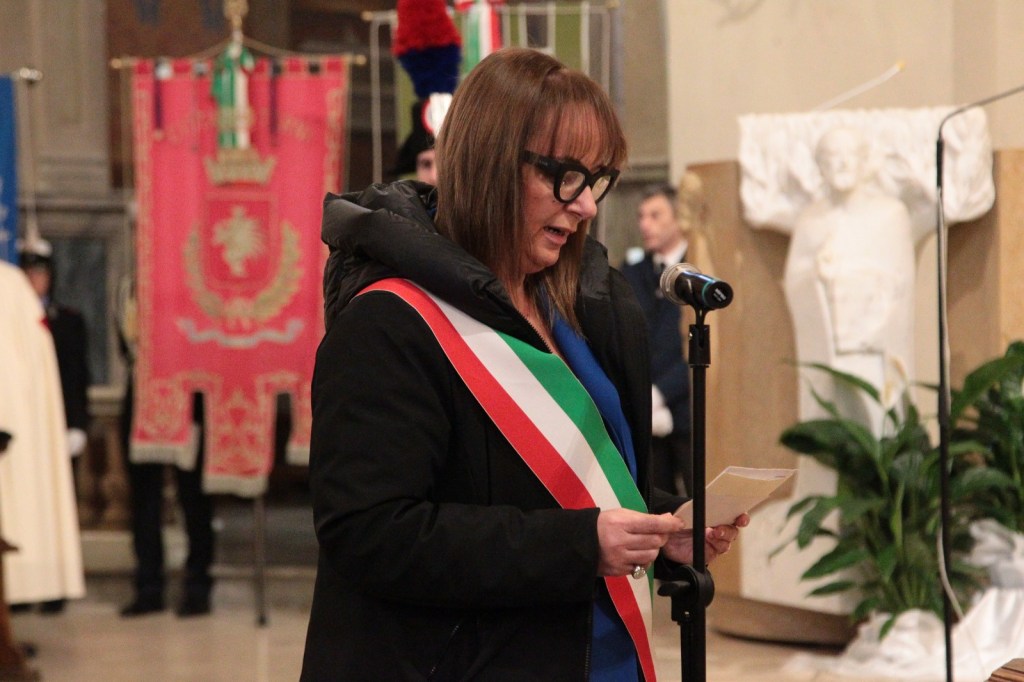 A female speaker wearing a ceremonial sash stands at a microphone holding a piece of paper, delivering a speech in a formal setting with decorative banners in the background.