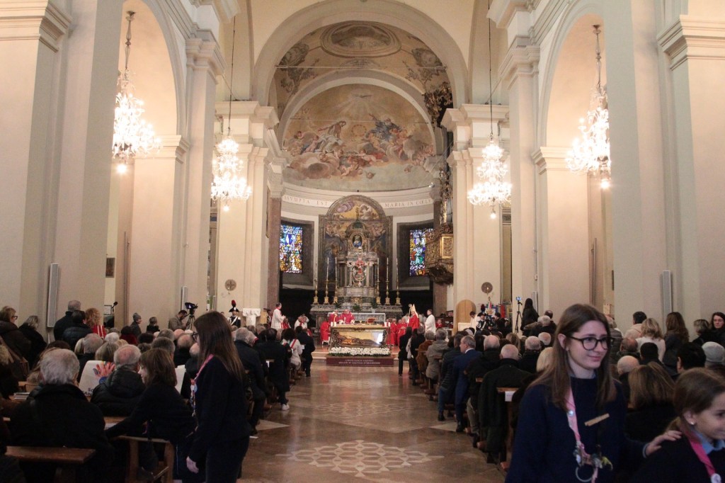 Interior view of a church filled with attendees during a ceremony, featuring ornate chandeliers and stained glass windows, with a procession at the altar.