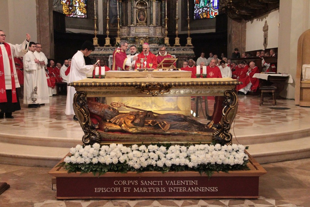 A religious ceremony taking place in a church, featuring clergy in red and white garments, gathered around an ornate altar. The focus is on a glass display containing the relics of Saint Valentine, surrounded by white flowers and ornate decorations.