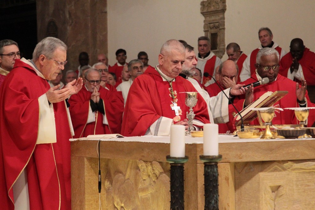 Clergy members in red robes participating in a religious ceremony, gathered around an altar with lit candles and communion vessels.
