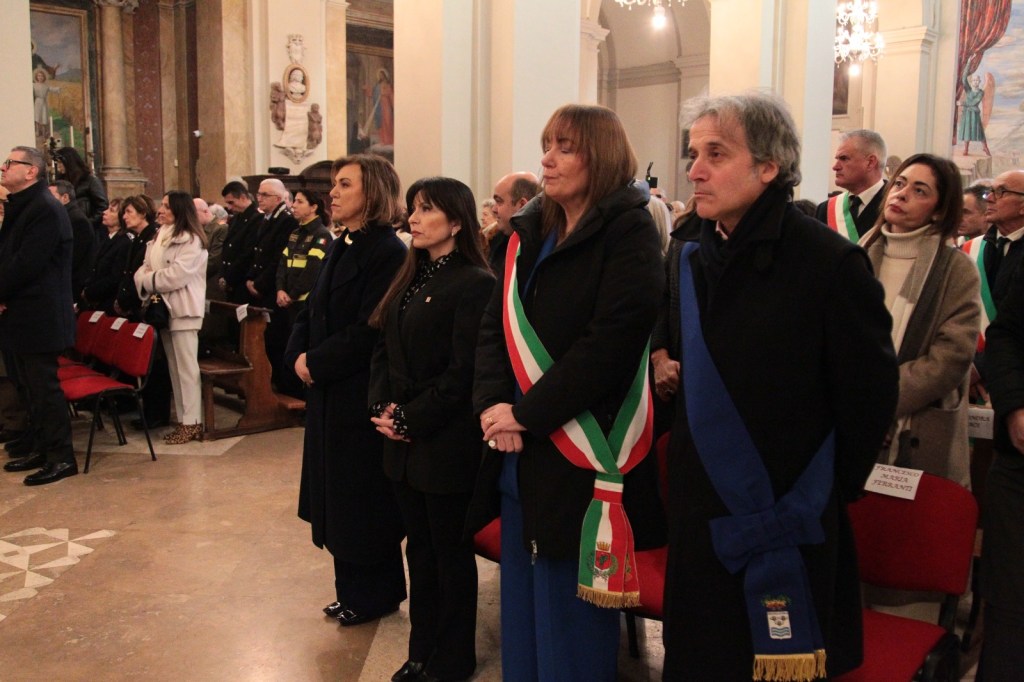 A group of people in formal attire standing in a church setting, some wearing ceremonial sashes. The scene appears to be part of an official event or ceremony.