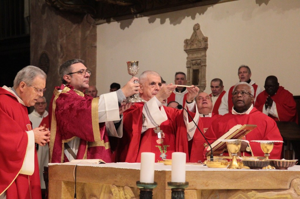 A group of priests in red vestments participating in a religious ceremony at the altar, with one priest holding a chalice and another displaying a host, surrounded by congregants and altar items.