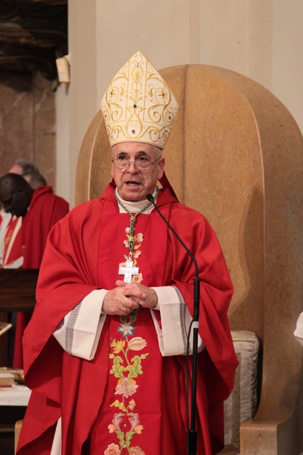 An elderly clergy member wearing a red vestment and a detailed, ornate mitre speaks into a microphone during a religious ceremony.