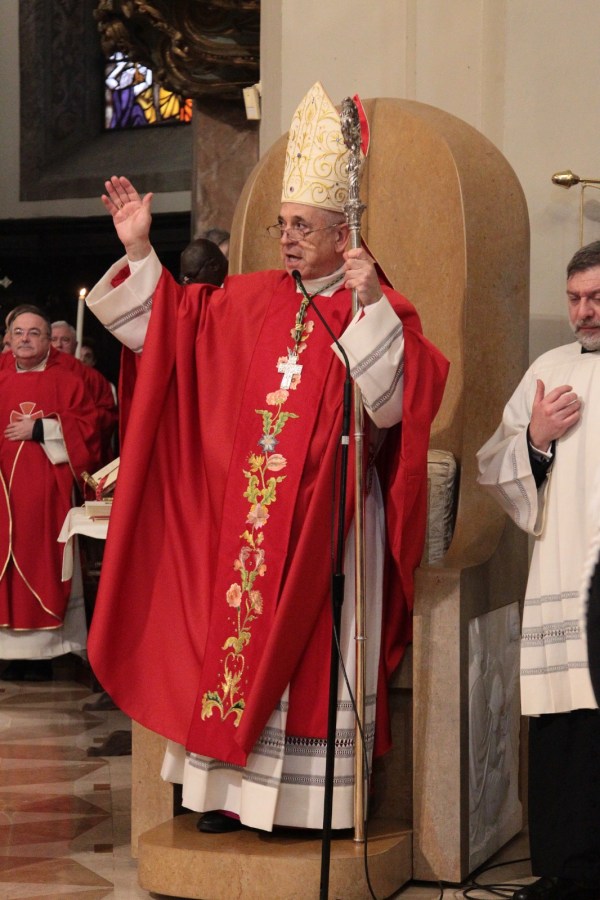 A religious leader in a red robe and ornate mitre gestures while holding a staff, surrounded by other clergy during a ceremony in a church.