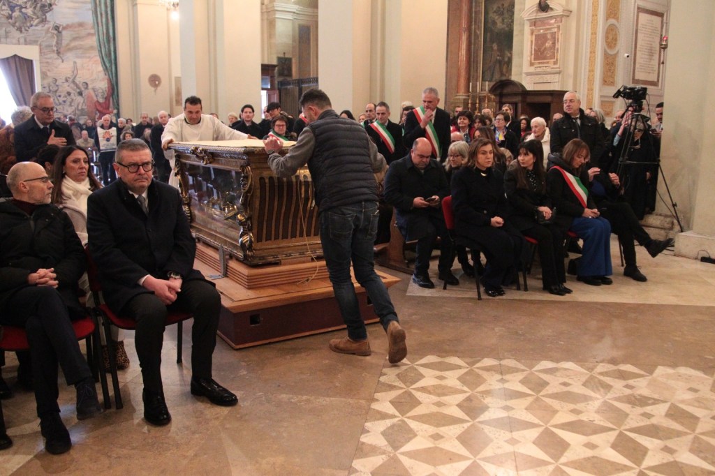 A gathering in a ceremonial setting with people seated and standing, observing a dignified event. A man in a vest is setting a decorative wooden structure, possibly a casket, while attendees, including officials in ceremonial sashes, watch intently.