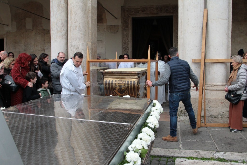 A funeral scene with mourners gathered around a wooden casket being carried out of a building. Some individuals are dressed in formal attire, while others are in casual clothing. Flowers adorn the foreground, and there are people looking on solemnly.