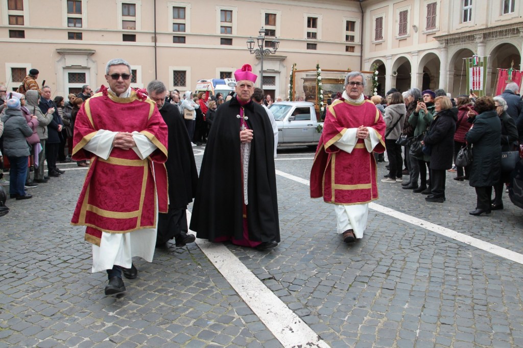 Clergy members in ceremonial robes walking in procession, with a crowd of people observing on the sides, in a street setting.