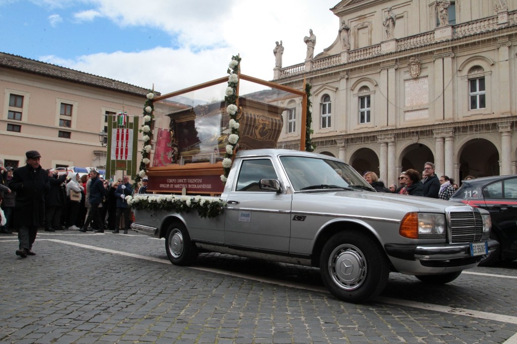A silver pickup truck adorned with flowers is driving in a procession, carrying a glass display that holds a religious artifact, with a crowd of onlookers and historic buildings in the background.