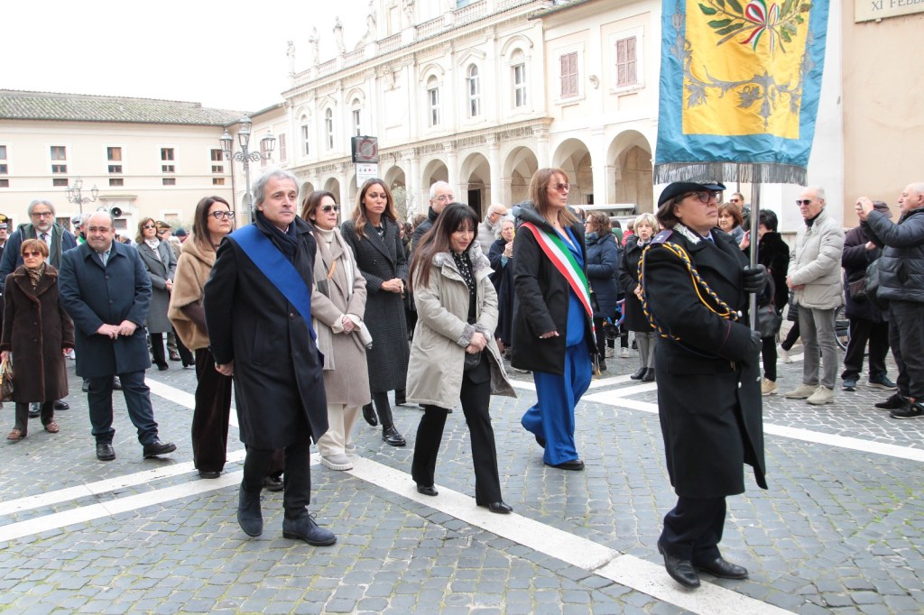 A group of people participating in a public event, walking in a square. In the foreground, a man in a suit and a sash leads the procession, while others follow, including a woman in formal attire and another carrying a flag. The background shows historical buildings and a crowd observing the event.