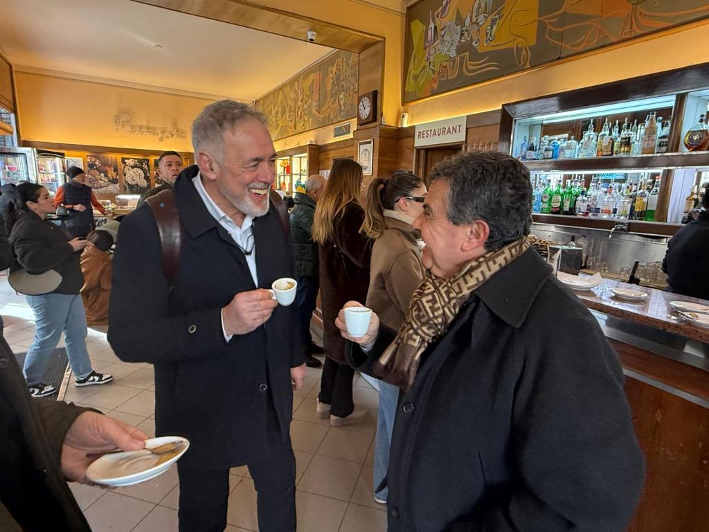 Two men laughing and conversing while holding cups of coffee in a busy café, with people in the background enjoying drinks and snacks.