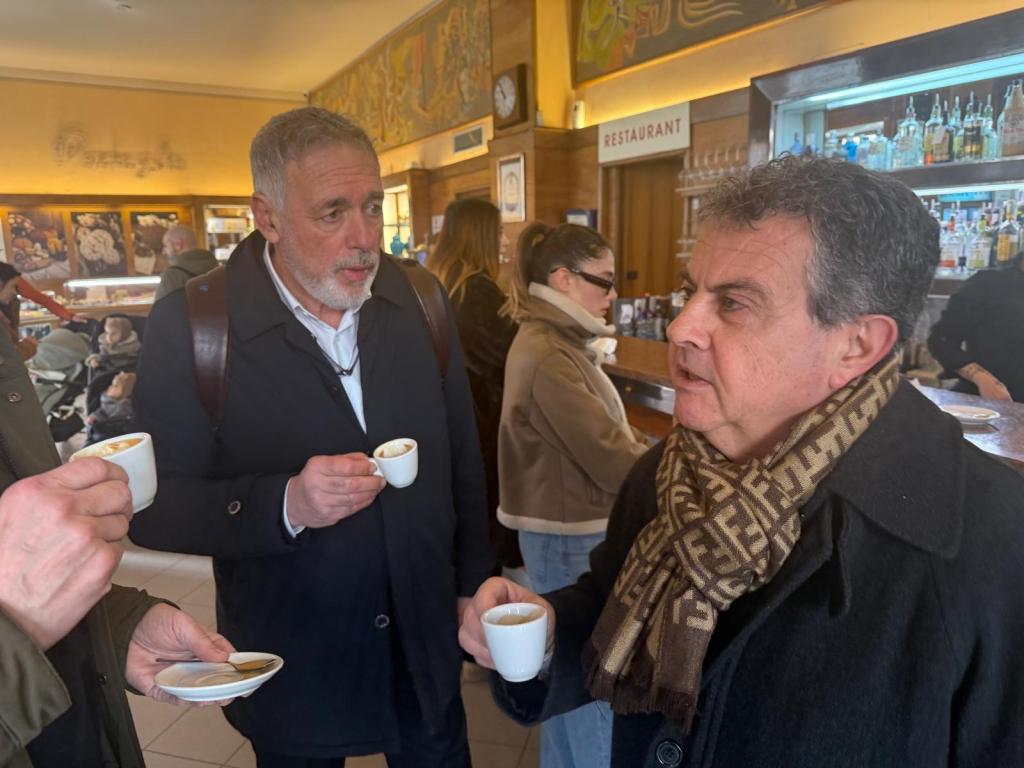Two men engaged in conversation while holding small cups of espresso in a bustling café setting.