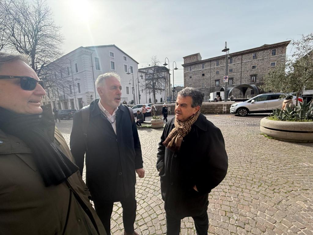 A group of three men engaged in conversation outdoors, with historic buildings and vehicles in the background under a clear sky.