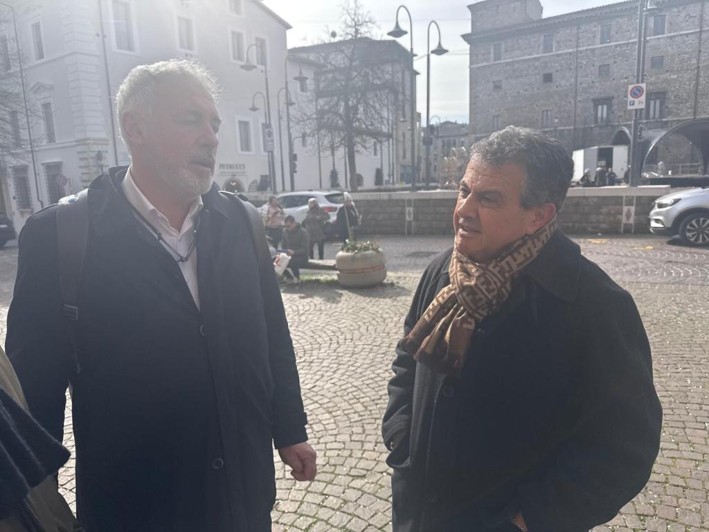 Two men conversing outdoors in a cobblestone area, with buildings and people in the background.