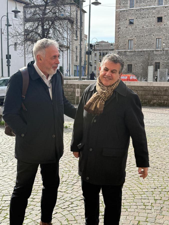 Two men in winter coats conversing outdoors, with a cityscape in the background featuring buildings and a tree.