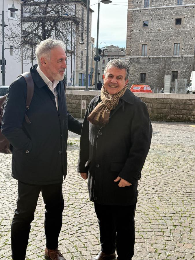 Two men standing on a cobblestone street, smiling and conversing, with buildings and trees in the background.