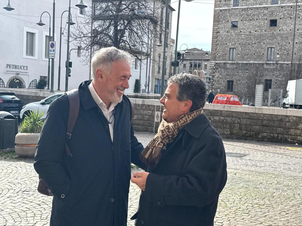 Two men engaged in a friendly conversation outdoors in a city setting, with buildings and parked cars in the background.