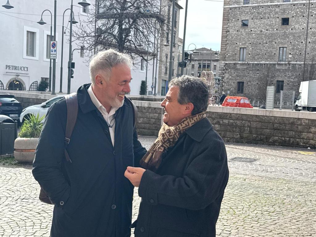 Two smiling men in conversation outdoors, with a historic building and parked cars in the background.