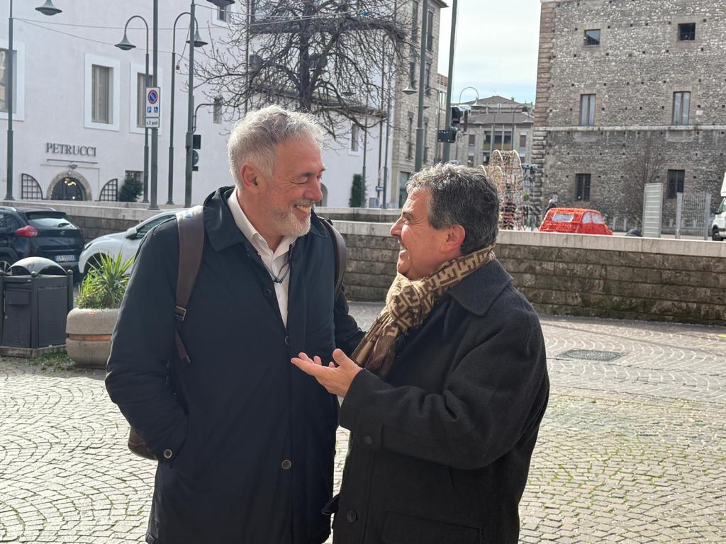 Two men engaged in a friendly conversation outdoors, smiling as they stand on a cobblestone street. In the background, there are buildings and parked cars.