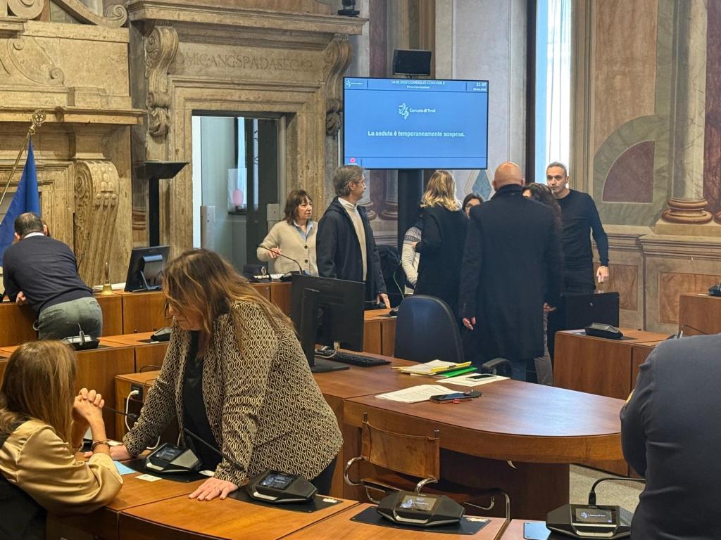 A meeting in a government chamber with individuals engaging in conversations. Desks with telephones and papers are present, alongside decorative walls and a digital screen displaying a message.
