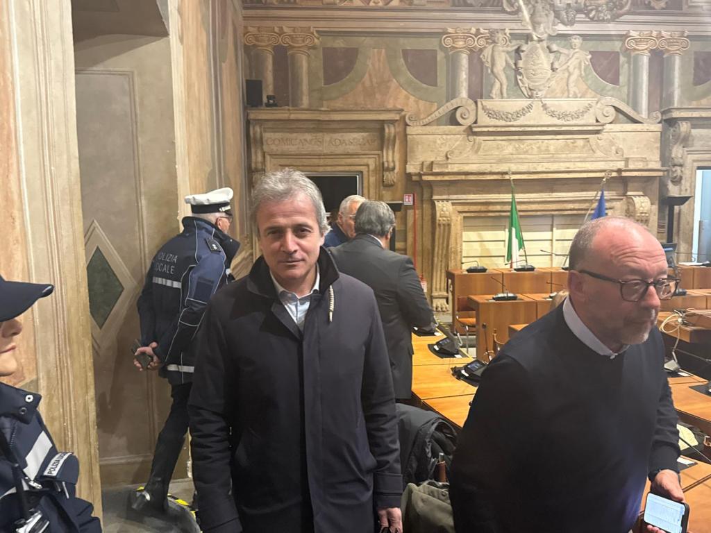 A group of people in a historic meeting room, with one man in the foreground wearing a dark coat and another in a black sweater. Police officers are visible in the background, standing near flags and a large decorative wall.