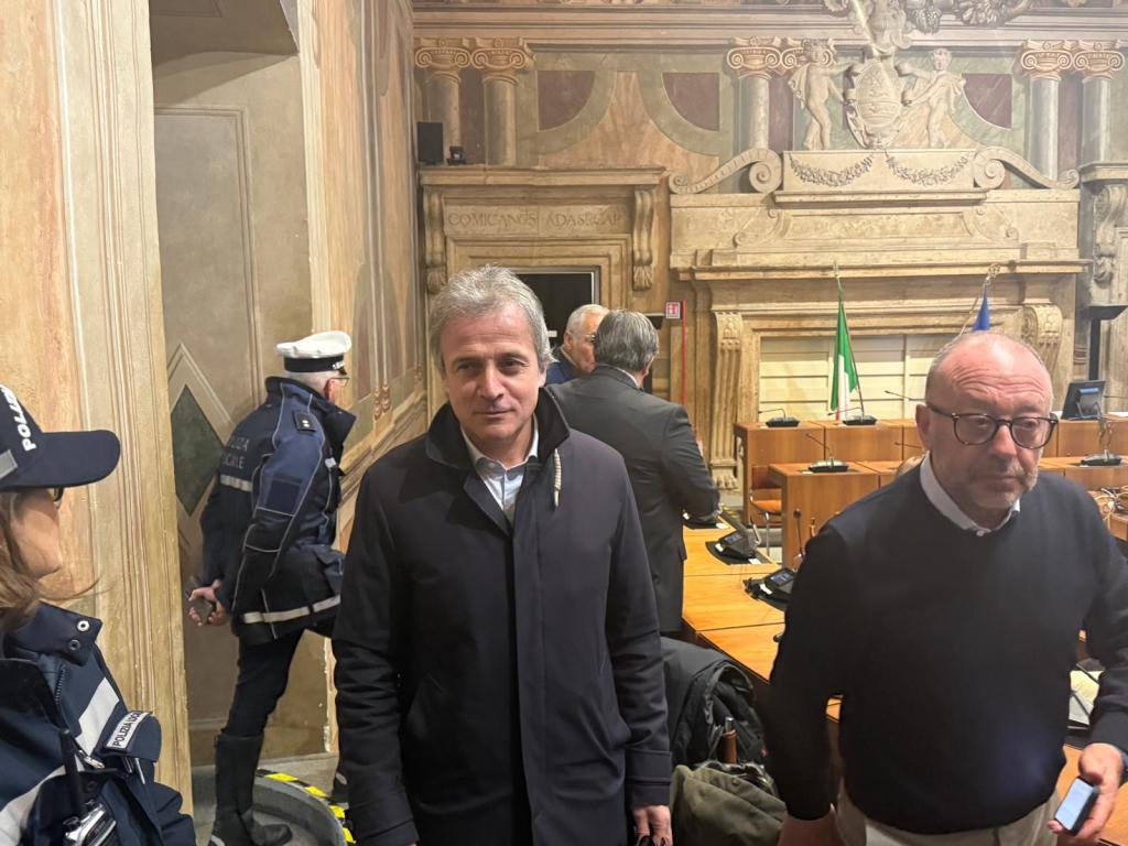 A gathering in a formal meeting room featuring several individuals, including a man in a dark coat and another man with glasses, amidst a historic setting with decorative walls and flags.