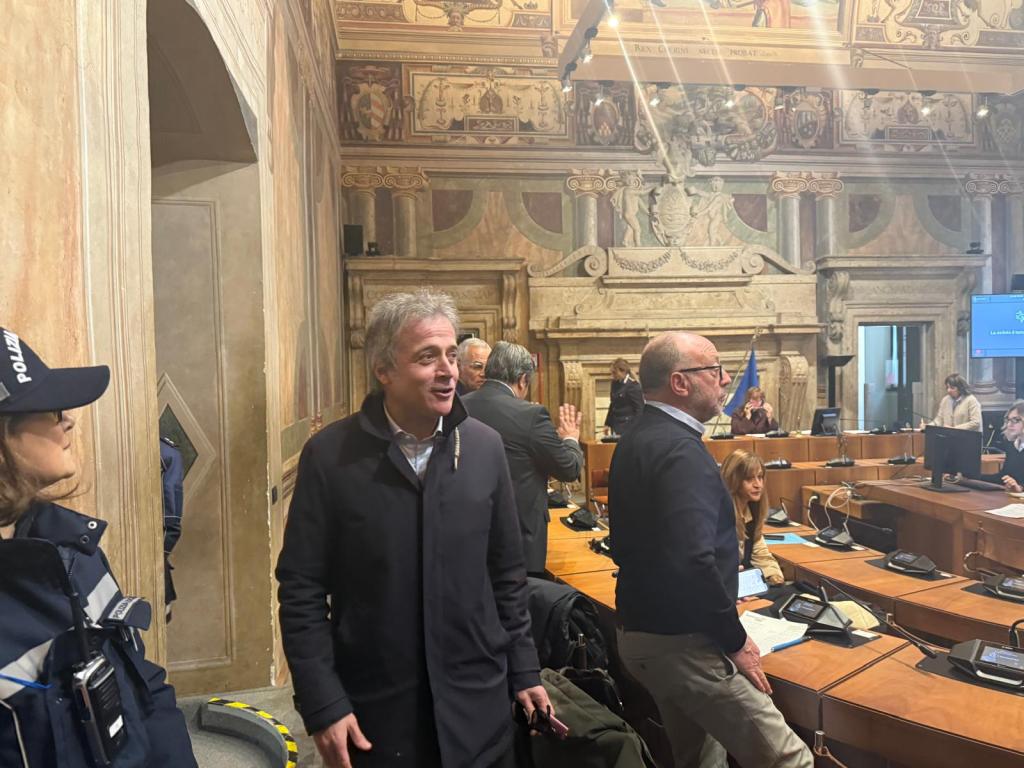 A group of people engaged in discussion inside a formal meeting room with intricate architectural details, including paintings and a decorative ceiling.