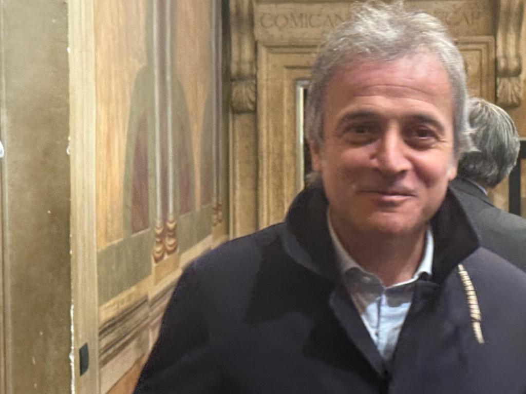 A smiling middle-aged man wearing a dark jacket, standing in front of a decorative wall with classical artwork.