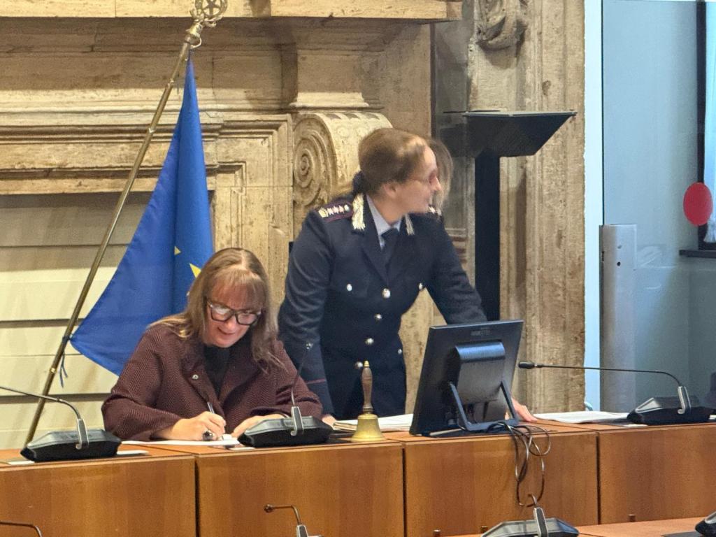 A woman in a dark coat writing at a table with a bell and two telephones, while a uniformed officer stands beside her in a meeting room with an EU flag in the background.