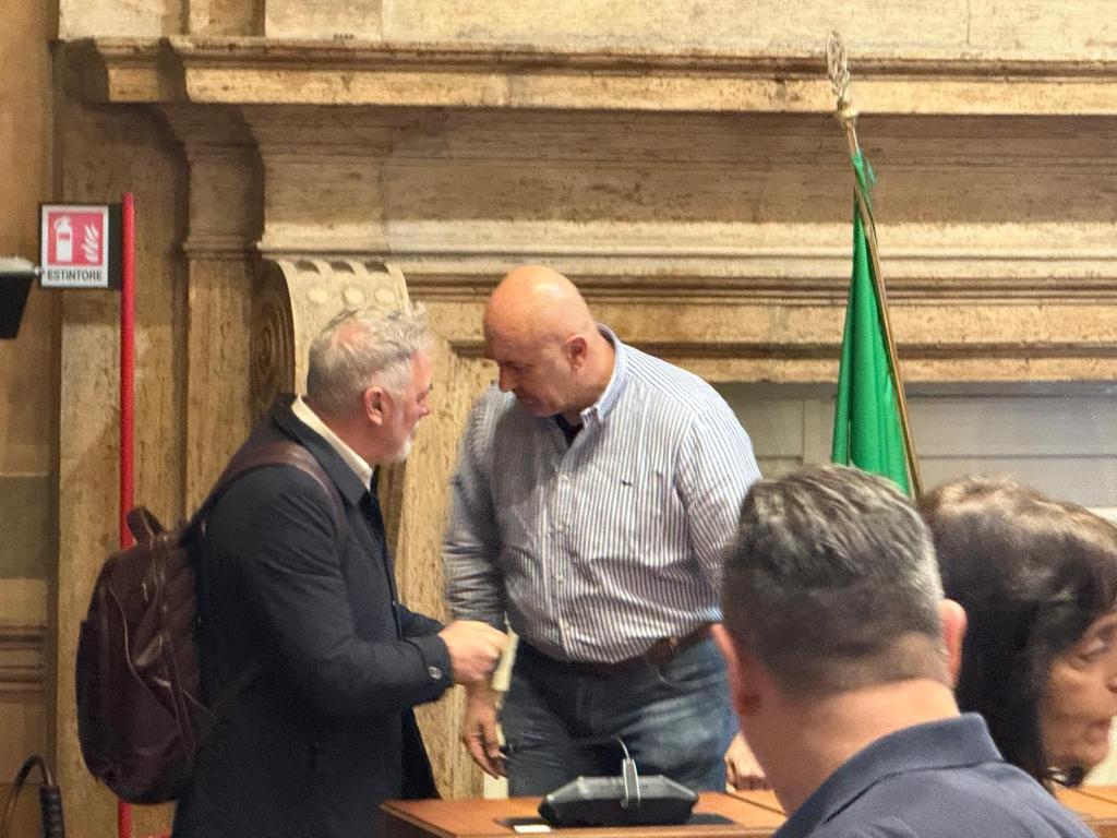 Two men engaged in conversation in a formal setting, with a green flag and ornate wall in the background.