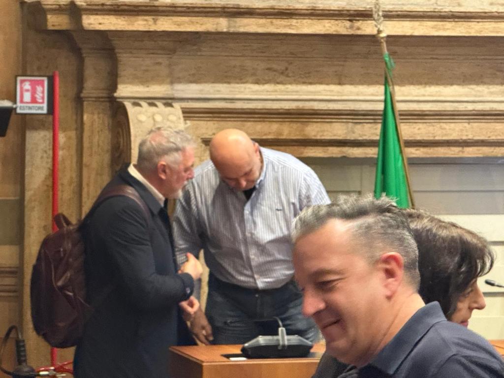 Two men engaged in conversation at a podium in a formal setting, with one man nodding and the other gesturing. A third man sits in the foreground, smiling.