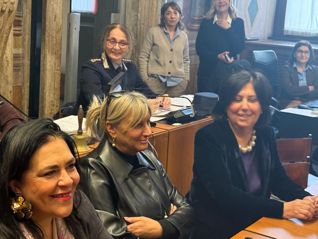 A group of women sitting at a table in a meeting room, smiling and engaged in conversation, with a few additional women standing in the background.