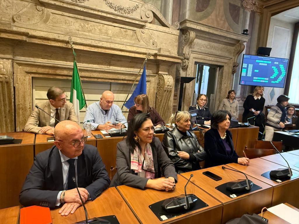 A group of people seated at a meeting table in a formal setting, with flags in the background and a digital screen displaying information.
