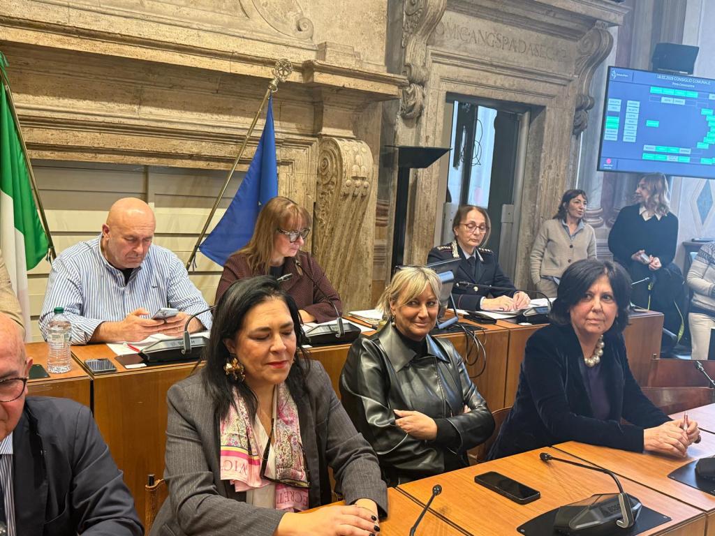 A group of people seated around a table in a formal setting, with flags in the background and a digital display showing information.