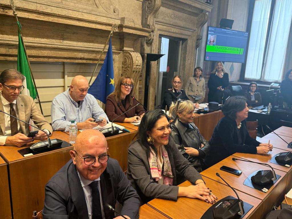 A group of people seated at a long wooden table in a formal meeting setting, with flags in the background and a screen displaying information.