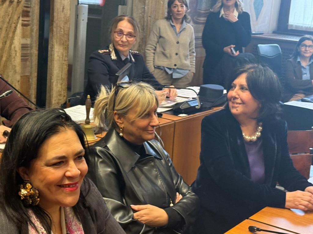 A group of women sitting at a table in a meeting room, smiling and engaged in conversation, with some women in the background observing.