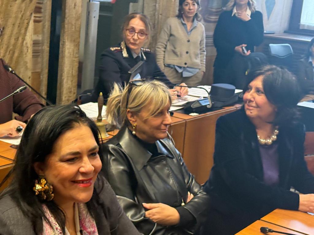 A group of women sitting at a conference table, engaged in conversation, with some participants visible in the background.