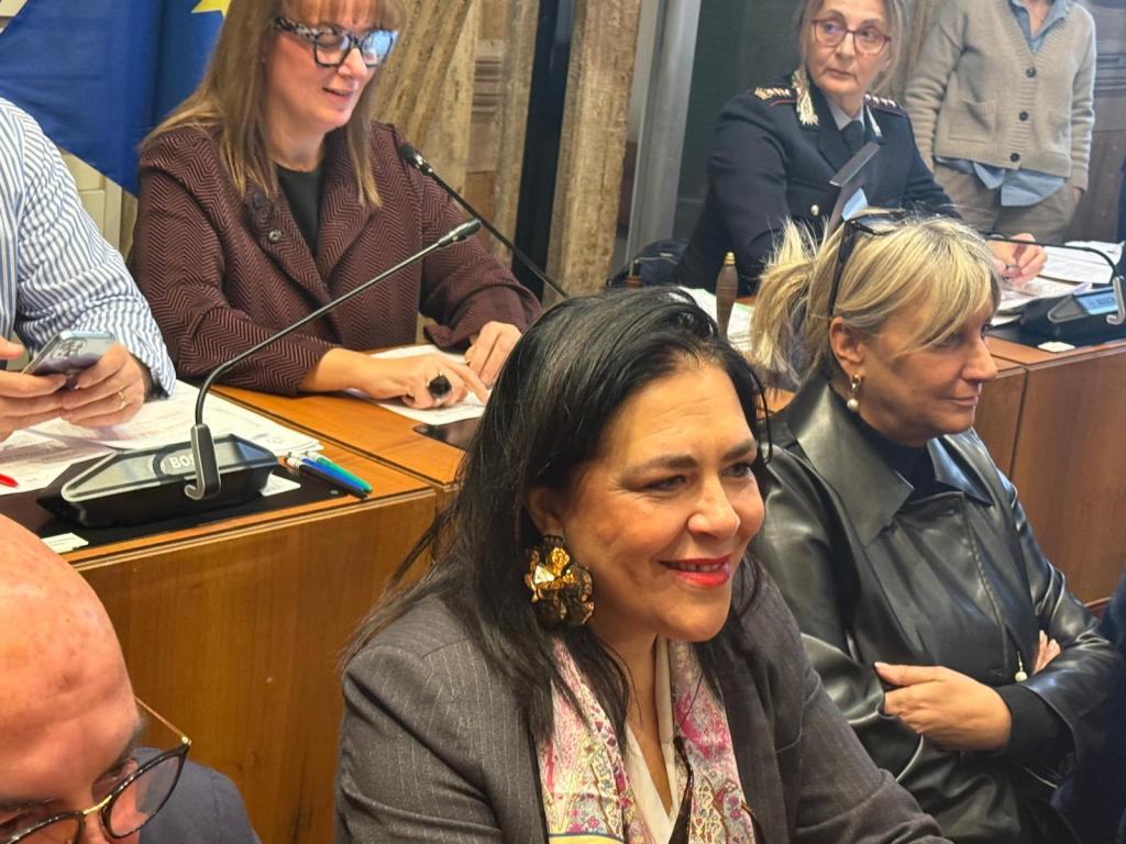 A group of people seated at a table during a meeting or discussion, with a woman in the foreground smiling and wearing large earrings.