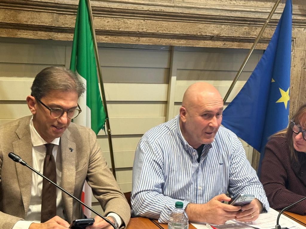 A meeting featuring three individuals seated at a table, with a green Italian flag and a blue European Union flag in the background. One person, wearing glasses and a beige suit, is looking at their phone while another, in a striped shirt, is speaking. A woman in a brown sweater appears to listen attentively.