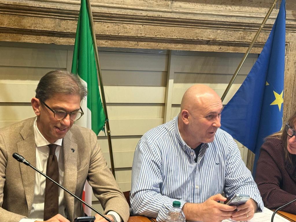Two men sitting at a table during a meeting, with the Italian and European Union flags in the background.