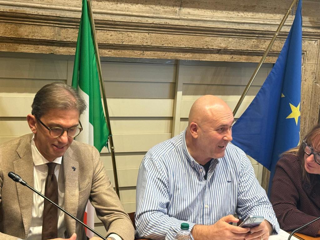 A meeting scene featuring three people seated at a table, with an Italian flag and a European Union flag in the background. One man is speaking while looking at his phone, another man is listening and smiling, and a woman is sitting next to them.