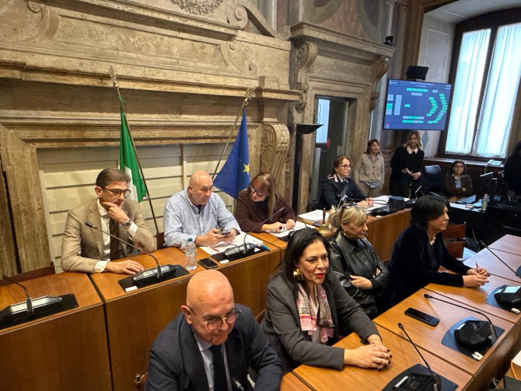 A group of officials seated at a conference table in a formal meeting room, with flags in the background and a large screen displaying information.